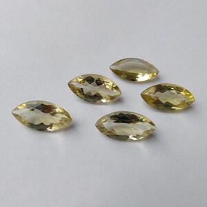 May include: Five marquise-cut gemstones with a pale yellow hue are arranged on a white surface. The gems have a faceted appearance, catching the light and displaying internal reflections. The overall composition suggests a collection of loose gemstones.