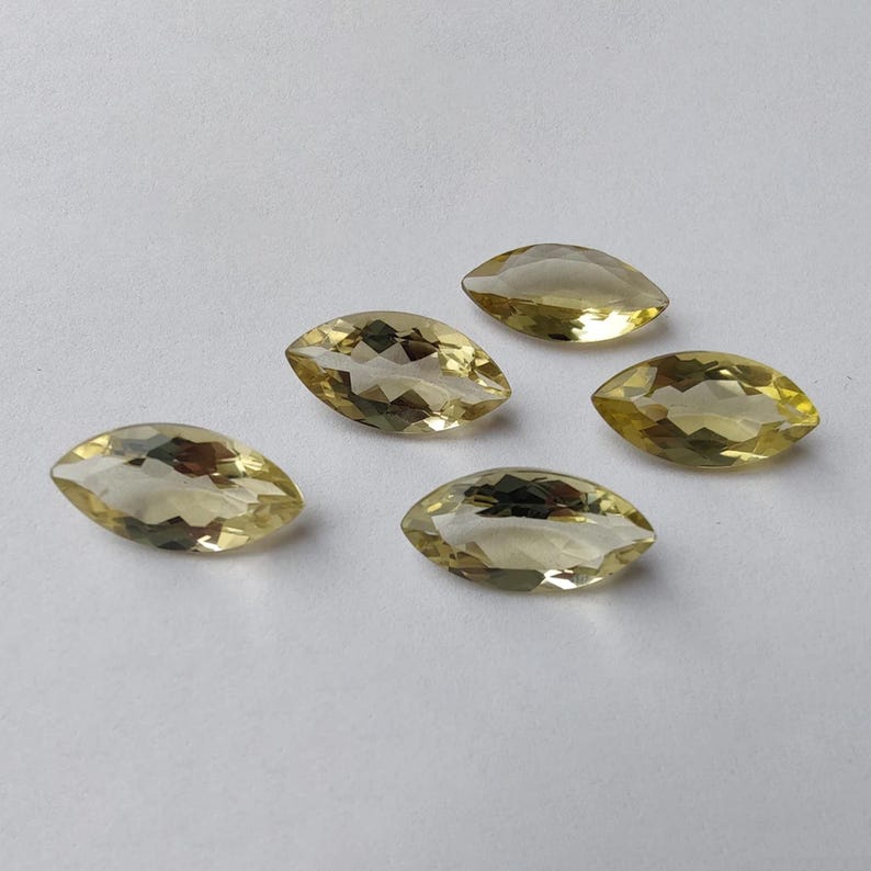 May include: Five marquise-cut gemstones with a pale yellow hue are arranged on a white surface. The gems have a faceted appearance, catching the light and displaying internal reflections. The overall composition suggests a collection of loose gemstones.