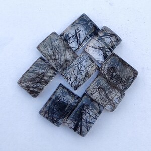 May include: A collection of rectangular gemstones with a clear base and intricate black needle-like inclusions. The stones are arranged in a slightly overlapping pattern, showcasing their unique internal structures. The overall effect is a display of natural mineral formations.