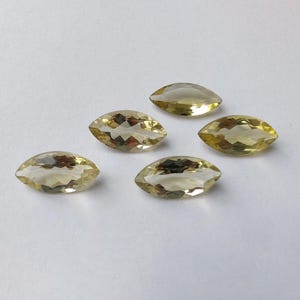 May include: Five marquise-cut gemstones with a pale yellow hue are arranged on a white surface. The faceted stones have a pointed oval shape, catching the light and displaying internal reflections. These gemstones are suitable for jewelry making.