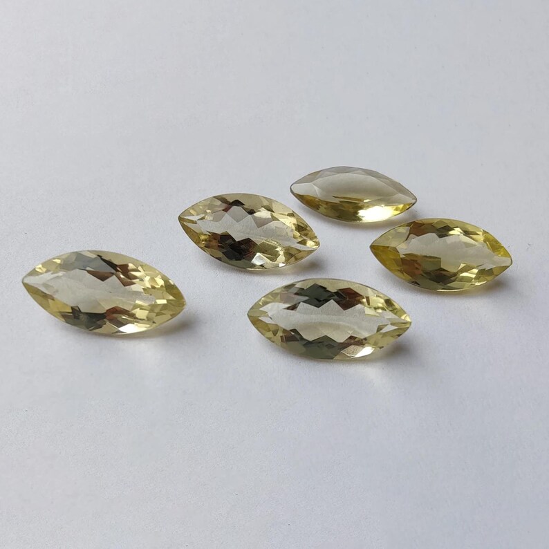 May include: Five marquise-cut gemstones with a pale yellow hue are arranged on a white surface. The gems have multiple facets that catch the light, creating a sparkling effect. These gemstones are suitable for jewelry making.