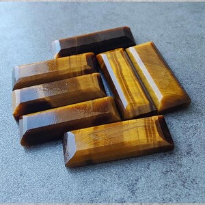 May include: A selection of tiger's eye gemstones, each cut into a rectangular shape. The stones are a warm, golden-brown colour, with bands of lighter and darker tones. The surfaces are polished and reflective.
