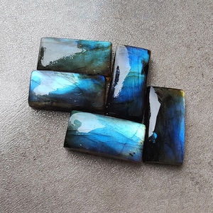 Pode incluir: Uma coleção de pedras retangulares de labradorita. As pedras exibem um vibrante jogo de cores, com azuis iridescentes, verdes e toques de ouro. Cada pedra tem uma superfície polida e brilhante, dispostas sobre um fundo neutro e texturizado.
