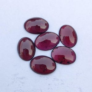 Op de afbeelding: Zes ovale, gepolijste edelstenen in een diepe bordeauxrode kleur. De stenen hebben een glad, reflecterend oppervlak, met een lichte variatie in tint. De edelstenen zijn in een cirkelvormig patroon op een witte achtergrond geplaatst.