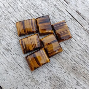 May include: Six rectangular tiger's eye gemstones with a polished, glossy finish. The stones display a rich, warm brown colour with natural golden-brown striations. The gemstones are arranged on a weathered wooden surface.