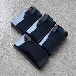 May include: Five rectangular, polished black stones arranged diagonally. Each stone has a glossy, reflective surface, with rounded edges. The stones are set against a light grey, textured background.