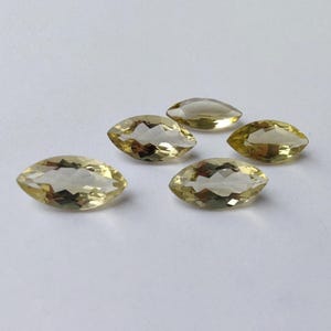 May include: Five marquise-cut gemstones with a pale yellow hue are arranged on a white surface. The gems have a faceted appearance, catching the light and displaying internal reflections. The overall composition suggests a collection of loose gemstones.