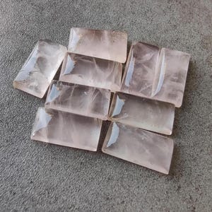 Könnte beinhalten: Eine Sammlung rechteckiger, durchscheinender Rosenquarzsteine. Die Steine haben ein poliertes, glasartiges Aussehen und variieren leicht in der Farbe, von blassrosa bis zu einem intensiveren Rosaton. Die Steine sind auf einer grauen Oberfläche angeordnet.