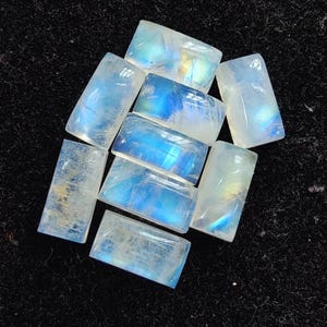 Könnte beinhalten: Eine Sammlung rechteckiger Mondstein-Edelsteine. Die Steine zeigen eine durchscheinende, milchig-weiße Basis mit schillernden blauen und gelben Reflexen. Die Edelsteine sind mit einer glatten, polierten Oberfläche geschliffen.