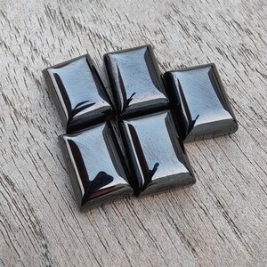 May include: Five polished, rectangular black gemstones on a light wooden surface. Each stone has rounded corners and a reflective, glossy finish. The gemstones are likely for jewellery or crafting, reflecting light.