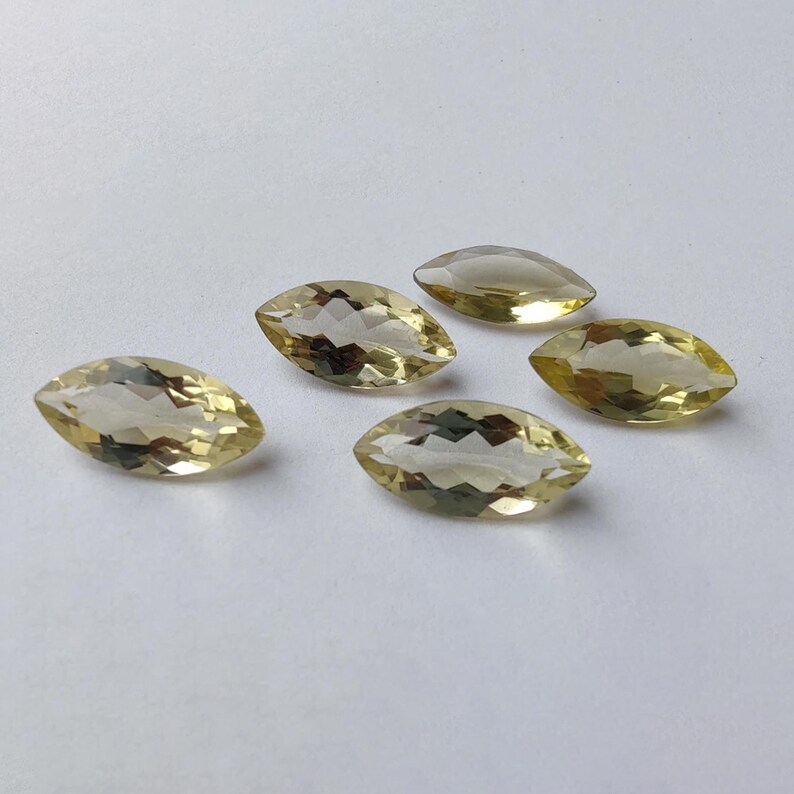 May include: Five marquise-cut gemstones with a pale yellow hue are arranged on a white surface. The gems have a faceted appearance, catching the light and displaying internal reflections. The stones are likely for jewelry making.