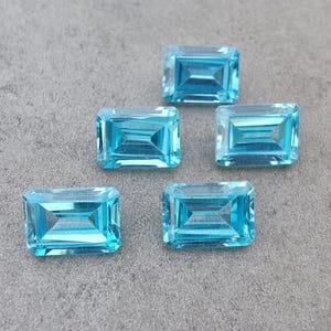 May include: Five rectangular, light blue gemstones with a faceted cut. The gems are arranged on a grey surface, showcasing their clarity and colour. The gems are a vibrant, transparent blue.