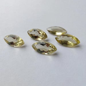 May include: Five marquise-cut gemstones with a pale yellow hue are scattered on a white surface. The gems have a faceted appearance, catching the light and displaying internal reflections. The elongated shape of each stone is prominent.