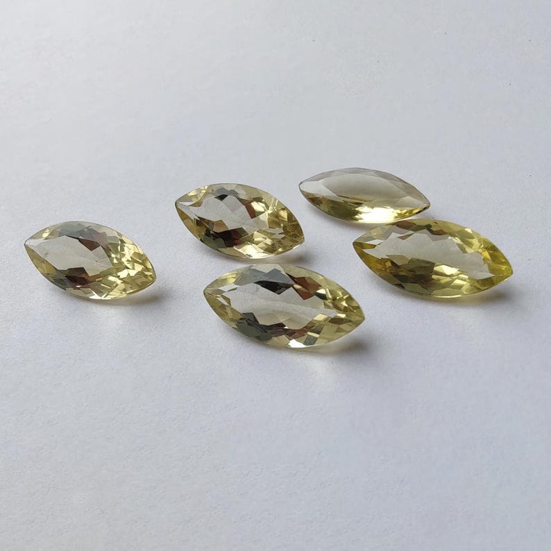 May include: Five marquise-cut gemstones with a pale yellow hue are scattered on a white surface. The gems have a faceted appearance, catching the light and displaying internal reflections. The elongated shape of each stone is prominent.