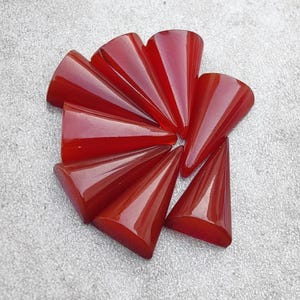 May include: A cluster of polished, cone-shaped gemstones in a vibrant, translucent red hue. The gemstones are arranged in a circular pattern, each with a smooth, glossy surface. The background is a light grey, textured surface.