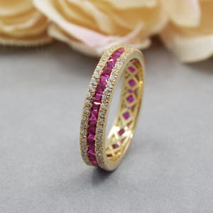 Pode incluir: Um anel de banda de ouro com uma dupla fila de pedras quadradas cor-de-rosa engastadas na banda. O anel é rodeado por pedras transparentes menores.