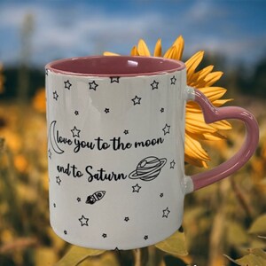 May include: White ceramic mug with a pink heart-shaped handle. The mug has a black and white design of stars, a crescent moon, a planet, and a rocket. The text on the mug reads "Love you to the moon and to Saturn".