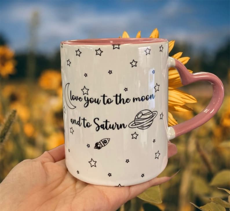 May include: White ceramic mug with a pink heart-shaped handle. The mug has a black and white design of stars, a crescent moon, a rocket, and the text "love you to the moon and to Saturn".