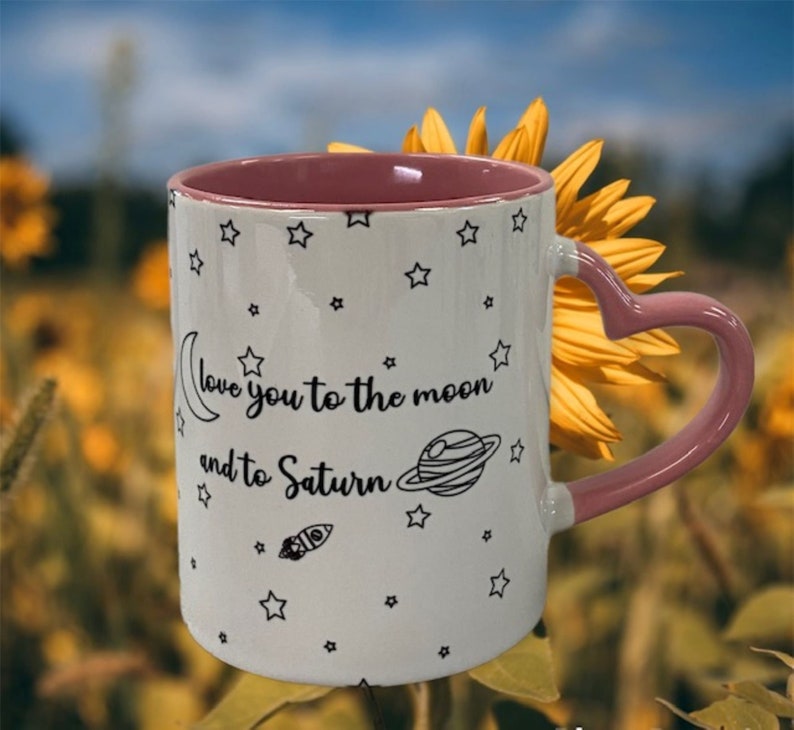 May include: White ceramic mug with a pink heart-shaped handle. The mug has a black and white design of stars, a crescent moon, a planet, and a rocket. The text on the mug reads "Love you to the moon and to Saturn".