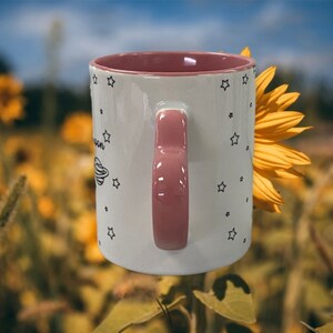 May include: White ceramic mug with a pink handle and a black and white design of stars and a planet. The mug is partially obscured by a yellow sunflower.