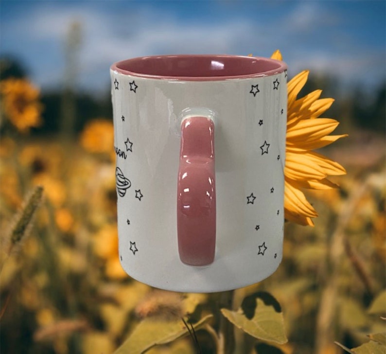 May include: White ceramic mug with a pink handle and a black and white design of stars and a planet. The mug is partially obscured by a yellow sunflower.