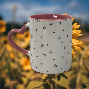 May include: A white ceramic mug with a pink heart-shaped handle and a black star pattern. The mug is partially obscured by a yellow sunflower.