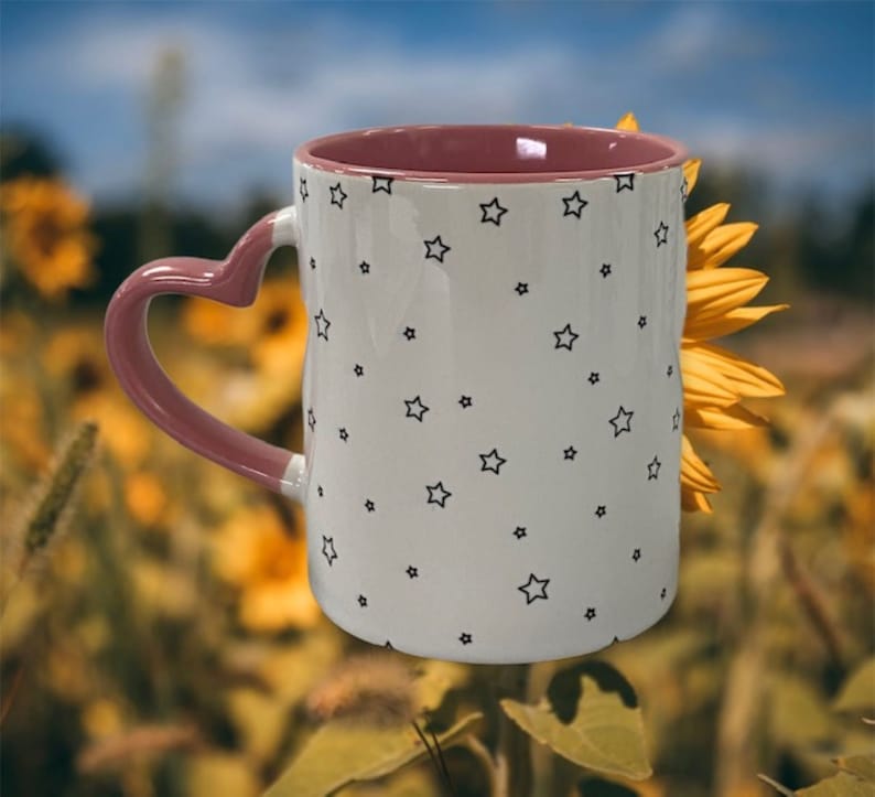 May include: A white ceramic mug with a pink heart-shaped handle and a black star pattern. The mug is partially obscured by a yellow sunflower.