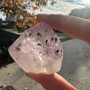 May include: A translucent, light purple crystal with dark inclusions is held between fingers. The crystal has a rough, natural shape and a clear, polished surface. The background shows a sunny outdoor scene with a tree and a sidewalk.