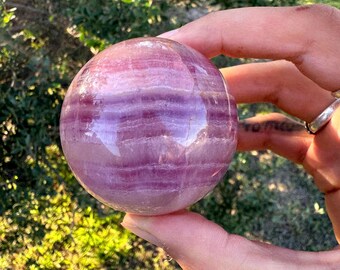 Op de afbeelding: Een gepolijste, bolvormige kristallen bol met banden van lavendel en paarse tinten. De bol wordt in een hand gehouden, met een vage groene achtergrond van bladeren en zonlicht.
