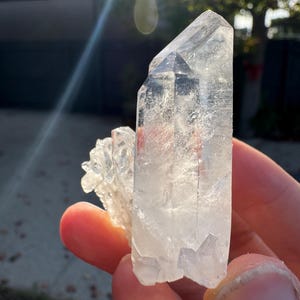 Op de afbeelding: Een heldere kwartskristalgroep. De belangrijkste kristal is groot en puntig, met kleinere kristallen eraan vast. Het kwarts is doorschijnend, met zichtbare interne structuren. Zonlicht benadrukt de helderheid van het kristal.
