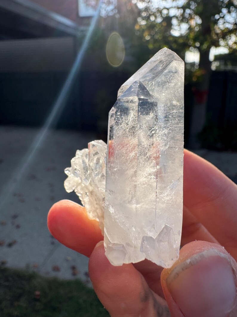 Op de afbeelding: Een heldere kwartskristalgroep. De belangrijkste kristal is groot en puntig, met kleinere kristallen eraan vast. Het kwarts is doorschijnend, met zichtbare interne structuren. Zonlicht benadrukt de helderheid van het kristal.