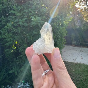 Op de afbeelding: Een heldere kwartskristalgroep met een groot, puntig centraal kristal en kleinere kristallen aan de basis. De kristal wordt in een hand gehouden, met een zilveren ring. De achtergrond toont groen gebladerte en zonlicht.