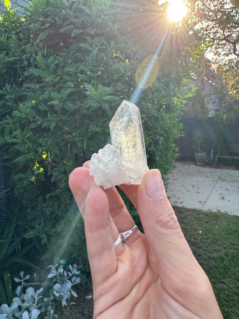 Op de afbeelding: Een heldere kwartskristalgroep met een groot, puntig centraal kristal en kleinere kristallen aan de basis. De kristal wordt in een hand gehouden, met een zilveren ring. De achtergrond toont groen gebladerte en zonlicht.