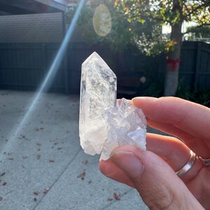 Op de afbeelding: Een heldere kwartskristalgroep met een groot, puntig kristal en kleinere, bevestigde kristallen. De kwarts wordt in een hand gehouden, met zonlicht op de achtergrond. Het kristal is transparant en lijkt natuurlijk.