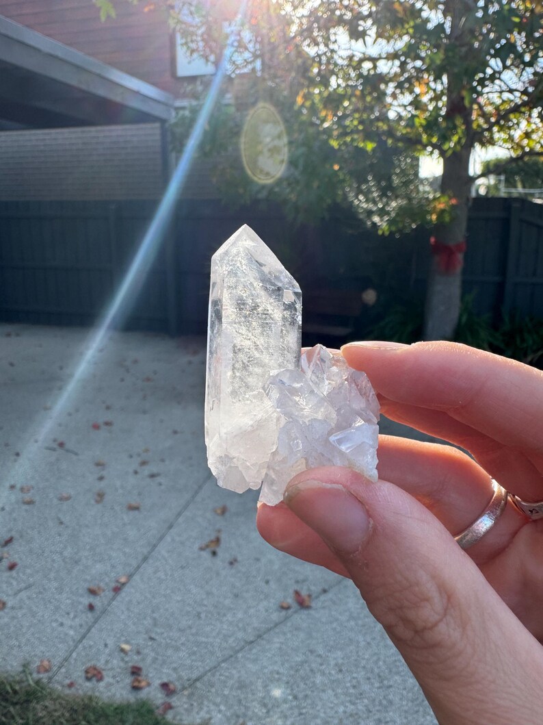 Op de afbeelding: Een heldere kwartskristalgroep met een groot, puntig kristal en kleinere, bevestigde kristallen. De kwarts wordt in een hand gehouden, met zonlicht op de achtergrond. Het kristal is transparant en lijkt natuurlijk.