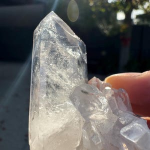Op de afbeelding: Een cluster van heldere kwartskristallen met een grote, puntige centrale kristal en kleinere kristallen eromheen. De kristallen zijn doorschijnend met een enigszins bewolkt uiterlijk. De foto is gemaakt in natuurlijk licht, met een heldere zonnevlam.