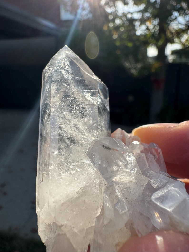 Op de afbeelding: Een cluster van heldere kwartskristallen met een grote, puntige centrale kristal en kleinere kristallen eromheen. De kristallen zijn doorschijnend met een enigszins bewolkt uiterlijk. De foto is gemaakt in natuurlijk licht, met een heldere zonnevlam.