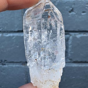 May include: A clear quartz crystal point with a natural, rough base. The crystal is held up against a dark gray brick wall. The quartz is transparent with visible internal structures and imperfections.