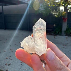 Op de afbeelding: Een heldere kwartskristalgroep met een groot, puntig centraal kristal en kleinere, bevestigde kristallen. Het kwarts wordt in een hand gehouden, waarbij zonlicht de facetten van het kristal verlicht. Het kristal heeft een bleke, doorschijnende kleur.