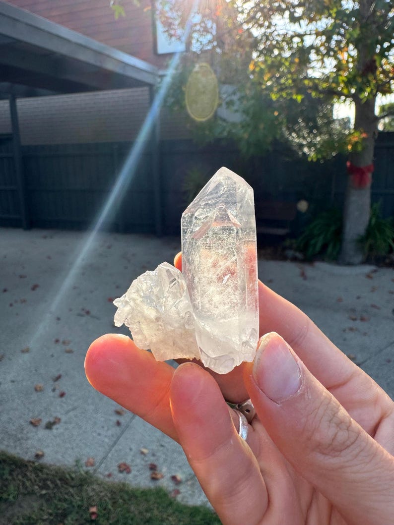 Op de afbeelding: Een heldere kwartskristalgroep met een groot, puntig centraal kristal en kleinere, bevestigde kristallen. Het kwarts wordt in een hand gehouden, waarbij zonlicht de facetten van het kristal verlicht. Het kristal heeft een bleke, doorschijnende kleur.