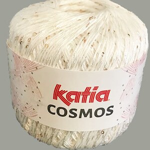 Könnte beinhalten: Ein Knäuel weißer Wolle mit goldenen Glitzerfäden. Die Wolle ist mit "Katia COSMOS" auf einem rosa-weißen Etikett gekennzeichnet.
