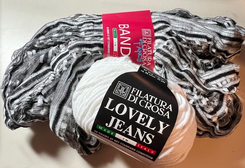 Pu&ograve; includere: Un gomitolo di lana bianco con un'etichetta nera che recita "Filatura Di Crosa Lovely Jeans Made in Italy 50 grammi in condizione standard netta". La lana &egrave; accanto a un tessuto a righe bianche e nere.