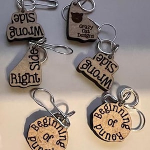 Puede incluir: Cinco marcadores de punto de madera con anillos de metal. Los marcadores están grabados con las palabras "Right Side", "Wrong Side", "Beginning of Round", "Wrong Side" y "Crazy Cat Designs".