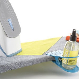 Puede incluir: Una tabla de planchar con una superficie gris, una plancha blanca y gris y un soporte para botella de spray azul con clip. El frasco de spray es transparente con una etiqueta amarilla que dice "Pineapple Grove". Un paño amarillo está en la tabla.