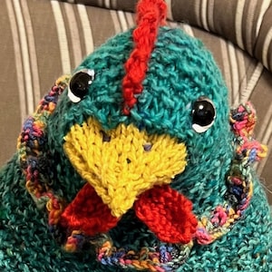 May include: Handmade knitted chicken toy with teal body, yellow beak, and red comb and wattle. The chicken has large black eyes and a colorful knitted collar. A whimsical and unique handcrafted item.