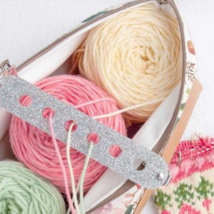 Puede incluir: Un estuche de tela con cremallera lleno de ovillos de lana en colores pastel, incluyendo rosa, amarillo y verde. Una guía de hilo plateada con purpurina y agujeros está colocada sobre la lana. Se ven agujas de tejer y un proyecto de tejido parcialmente terminado.