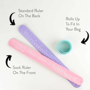 May include: A pink and lilac sock ruler with a rolled-up teal band. The pink ruler is labelled "Sock Ruler" and has measurements for kids, women, and men. The lilac ruler is labelled "Standard Ruler" on the back. The text says "Rolls Up To Fit In Your Bag."