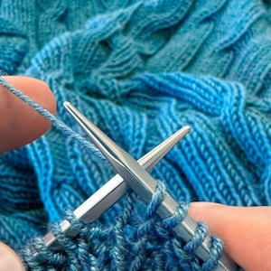 May include: Close-up of knitting needles with blue yarn. The needles are silver and are in the process of creating a knitted fabric. The yarn is a vibrant shade of blue, and the texture of the knitted fabric is visible.