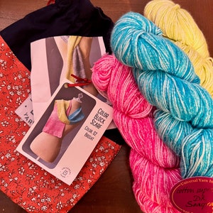 May include: A knitting kit for a colour block scarf with three skeins of yarn in pink, blue, and yellow. The yarn is labelled "Universal Yarn in cotton supreme Dk Seascape". The kit also includes a project bag and instructions.