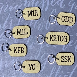 May include: Seven wooden tags with metal rings, each with a different knitting abbreviation: M1R, GDD, M1L, K2TOG, KFB, SSK, YO.